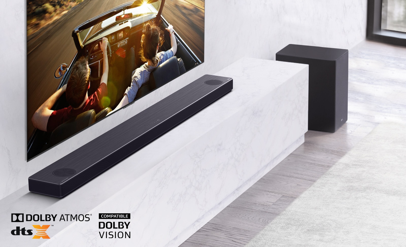 TV is on the wall, LG Soundbar is below on a white marble shelf with a sub-woofer to the right. TV shows a couple in a car. 