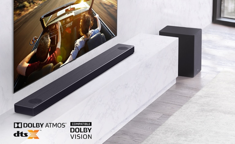 TV er på veggen, LG-lydplanken er under på en hvit marmorhylle med en subwoofer til høyre. TVen viser et par i en bil. 