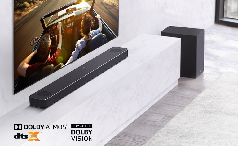 TV er på veggen, LG-lydplanken er under på en hvit marmorhylle med en subwoofer til høyre. TVen viser et par i en bil. 