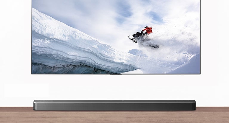 TV og lydplanke vist forfra. TV viser en mann som rir på en snøskuter i snøkledde fjell. HDMI 2.1-logoen vises under TVen. 