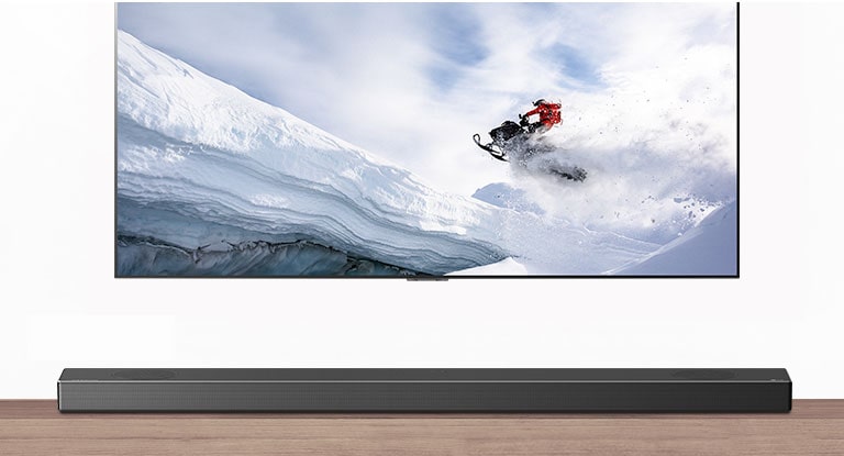 TV og lydplanke vist forfra. TV viser en mann som rir på en snøskuter i snøkledde fjell. HDMI 2.1-logoen vises under TVen. 