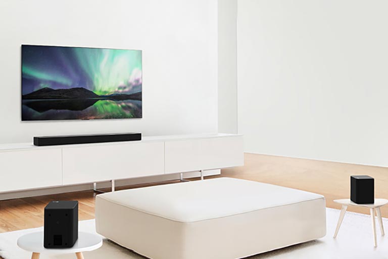 TV og lydplanke i en hvit stue med en hvit sofa i midten. Høyttalere sitter på begge sofa-endene.