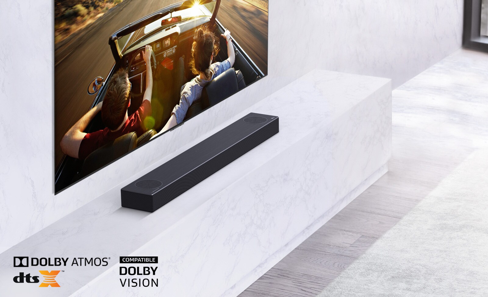 TV er på veggen, LG-lydplanken er under på en hvit marmorhylle med en subwoofer til høyre. TVen viser et par i en bil. 