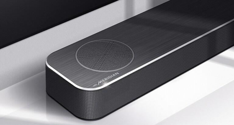Nærbilde av LG Soundbars venstre side med Meridian-logoen vist på nedre venstre hjørne av produktet. Nedre venstre side av TVen er også synlig. 