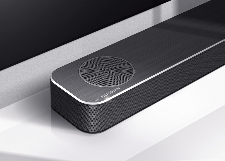Nærbilde av LG Soundbars venstre side med Meridian-logoen vist på nedre venstre hjørne av produktet. Nedre venstre side av TVen er også synlig.