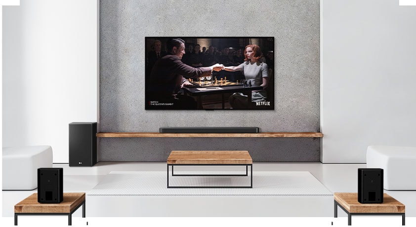 Et sett med to bakhøyttalere, subwoofer og en lydplanke og TV i en hvit stue.  En kvinne og en mann spiller sjakk på en TV-skjerm.