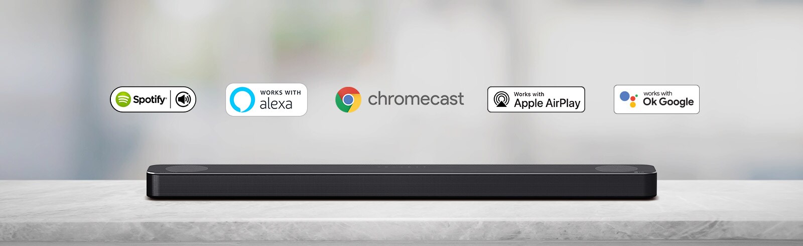 Det er plassert en lydplanke på en grå hylle og det er AI plattform-logoer, og logoene Spotify, Alexa, Chromecast, Apple Airplay og deretter OK Google kommer inn fra venstre mot høyre.