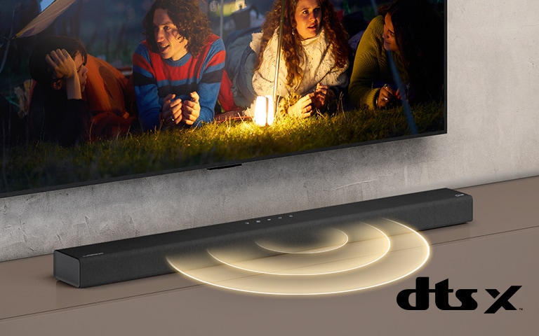 LG-TV-en henger på veggen. På skjermen vises to par som ligger i gresset. Foran dem står det en lampe. LG-lydplanken står under LG TV-en. Det kommer lydgrafikk ut fra fronten av lydplanken. DTS Virtual:X-logoen visen nederst i høyre hjørne av bildet.