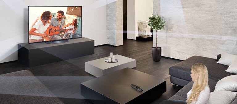 Et bredt bilde av et lyst rom med en TV opphengt på en vegg og en dame plassert på en sofa på motsatt vegg som ser på TV.