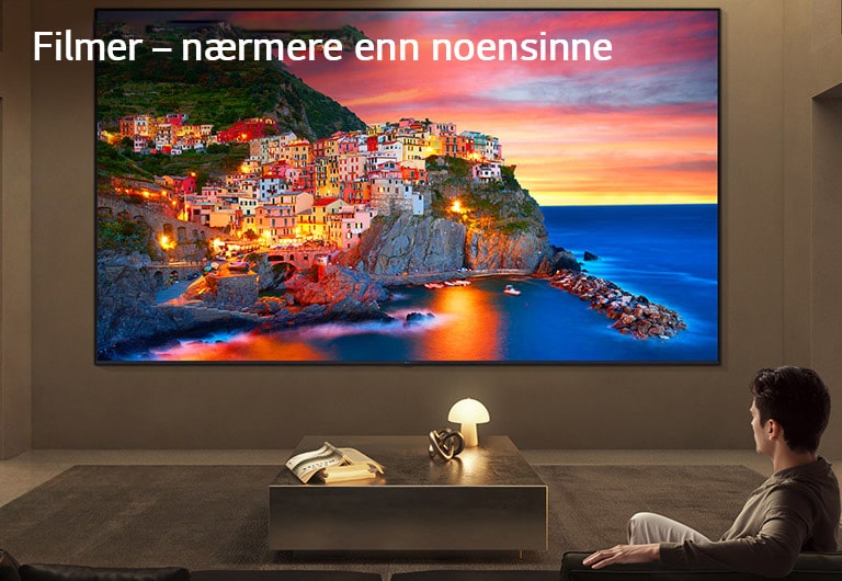 LG Ultra Big TV er veggmontert i en romslig stue, der en stor skjerm viser en levende kystlandsby ved solnedgang og illustrerer hvordan en større skjerm gir en oppslukende og filmatisk seeropplevelse.