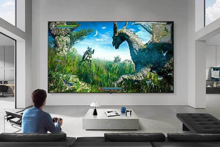 LG TV for gamere er montert på en vegg i en svart-hvitt-tonet stue og viser en fantasidinosaurspillscene på en stor skjerm, mens en person sitter i en sofa og holder en kontroller.