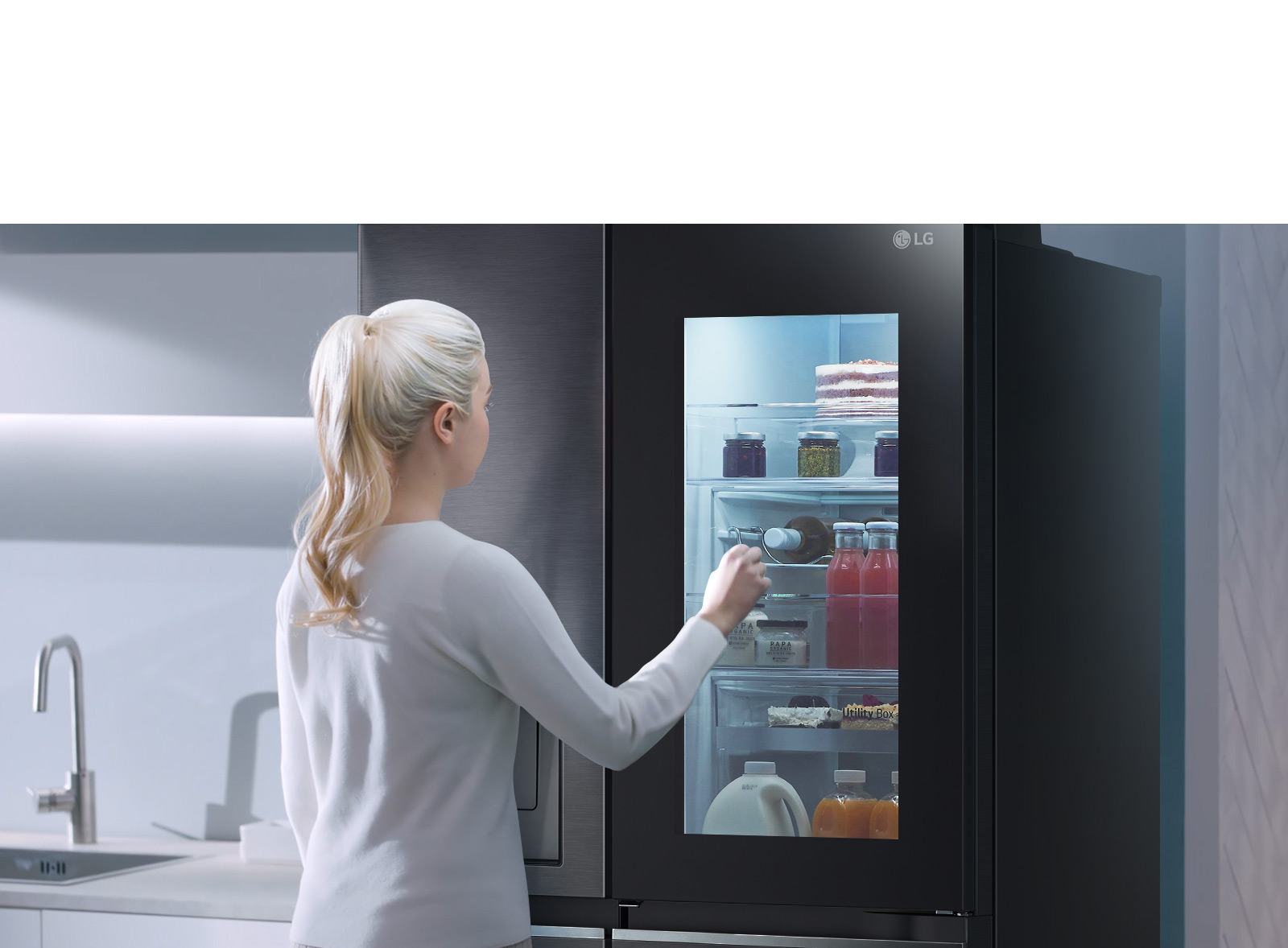 A video shows a woman approach her InstaView refrigerator and knock twice. The interior lights up and she can see the contents of her fridge without opening the door. The view zooms in to focus on the drinks in the door and then zooms out to see the woman from behind as she opens the door and grabs a drink.