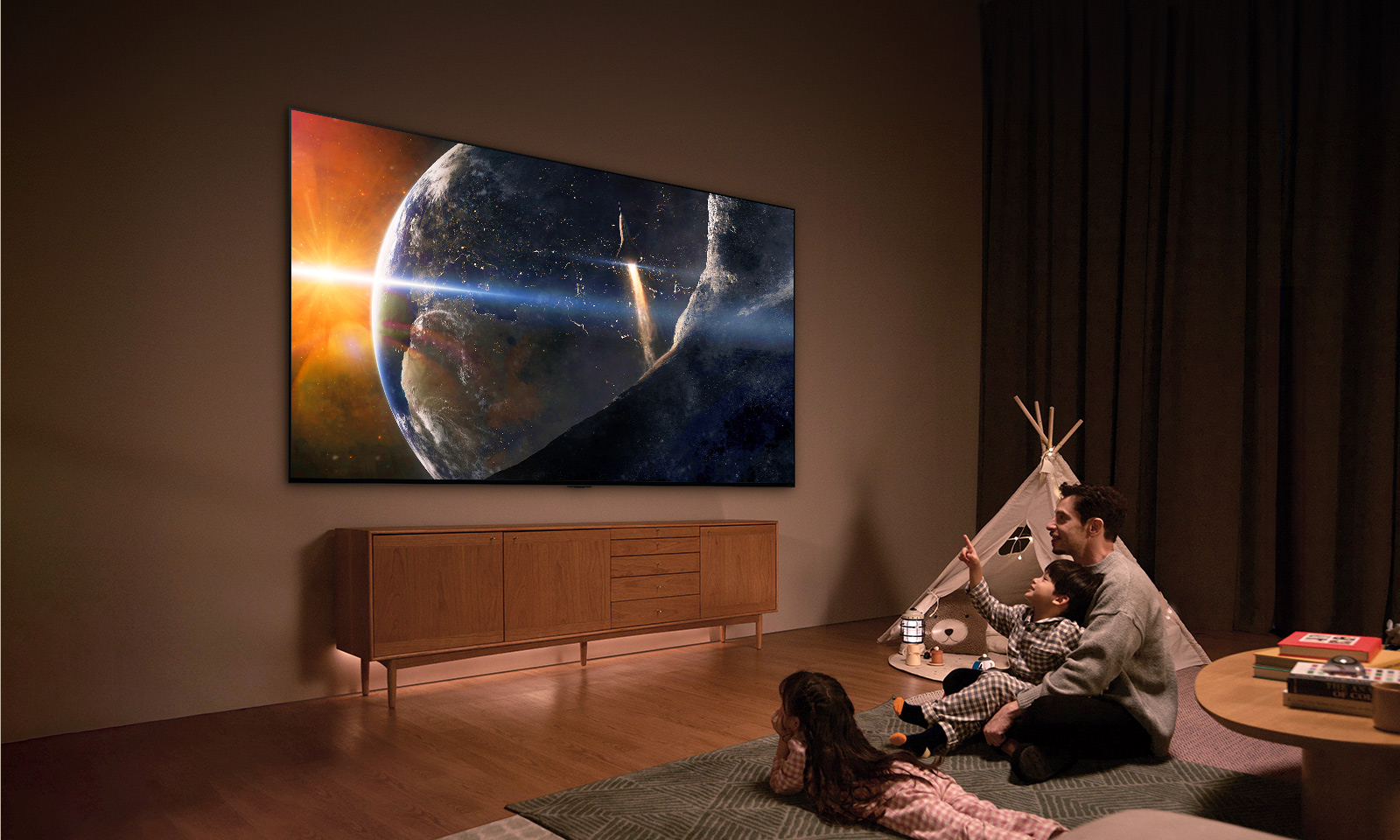 An image of a family sat on the floor of a low-lit living room by a small table, looking up at an LG TV mounted on the wall showing the Earth from space.