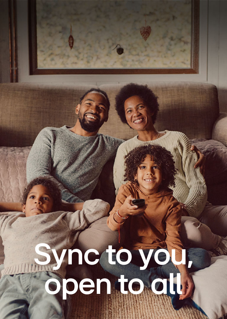 An image of a family of four sitting on the floor in front of cozy brown sofa looking straight ahead and laughing while watching TV. The words "Syncs to you, open to all" overlay the image in a white font. 