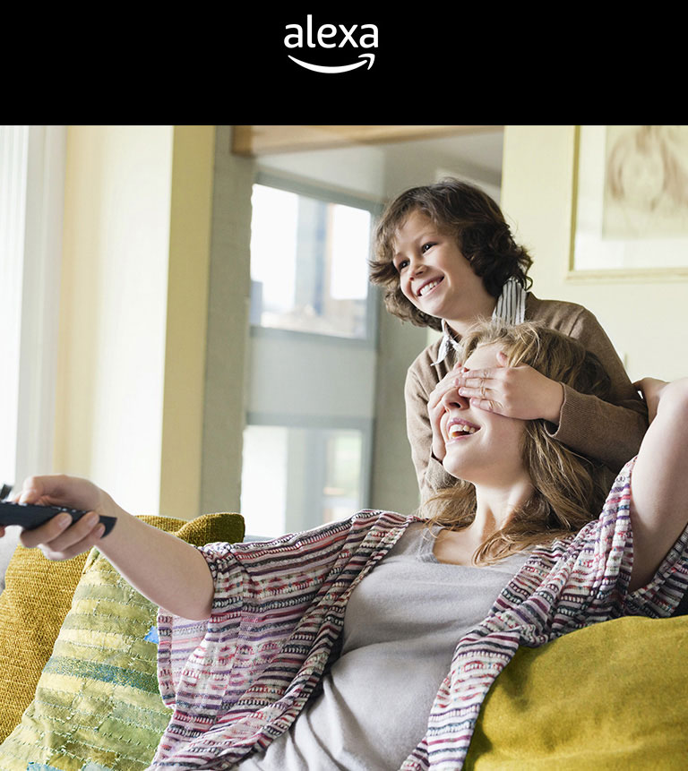 A kid is covering his mother’s eyes who is holding a TV remote. There is Google Assistant logo and alexa logo above image.