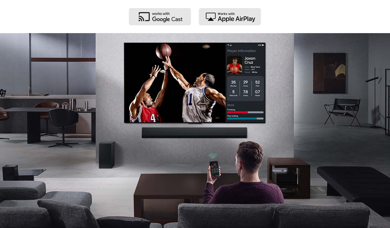 Person in their living room holding their phone. On the phone is a transmitting icon showing that the phone screen is being mirrored on the TV. On the TV is a basketball game to the side is the mirrored screen, showing player statistics.