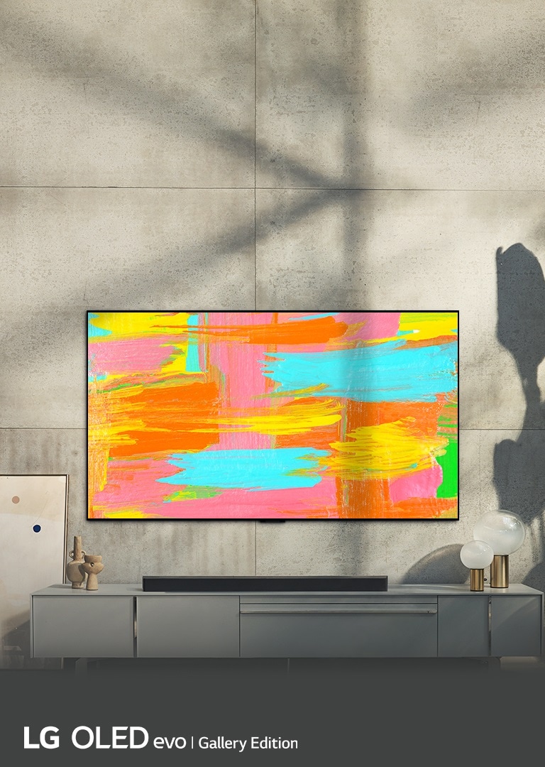 An image of LG OLED G2 hung on the wall of a minimalist gray room with a bright and vibrant abstract artwork displayed on the screen. The words "Make life a masterpiece" are displayed over the image.