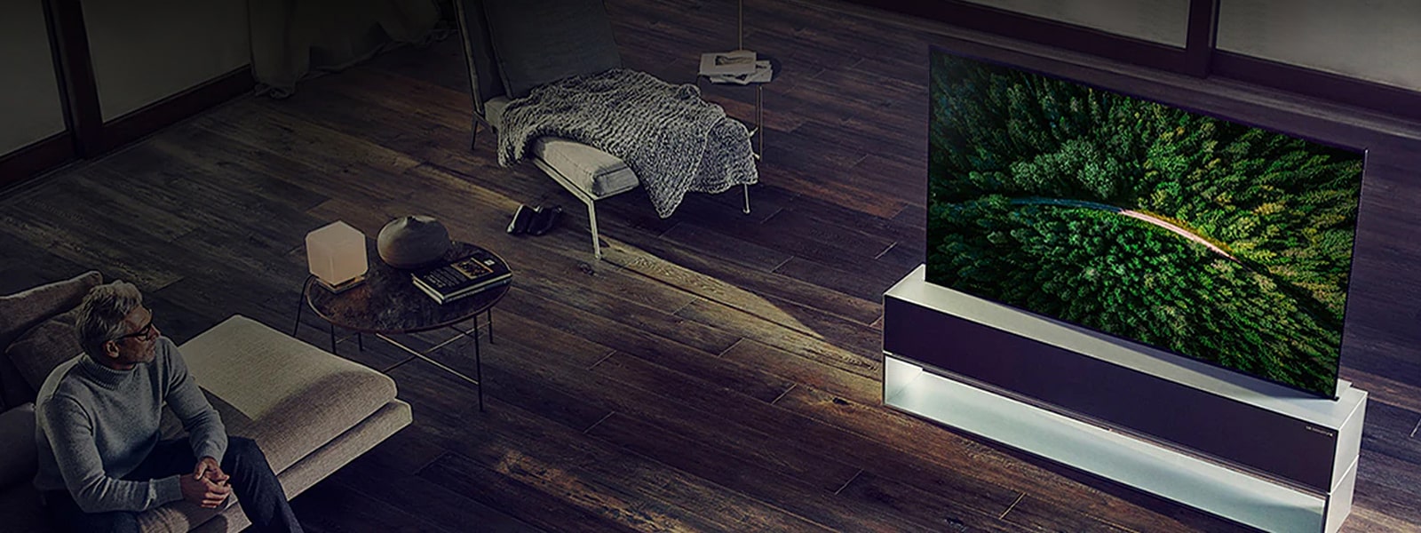 Telewizor LG SIGNATURE Rollable OLED TV jest umieszczony na drewnianej podłodze w luksusowym salonie, a mężczyzna ogląda telewizję.