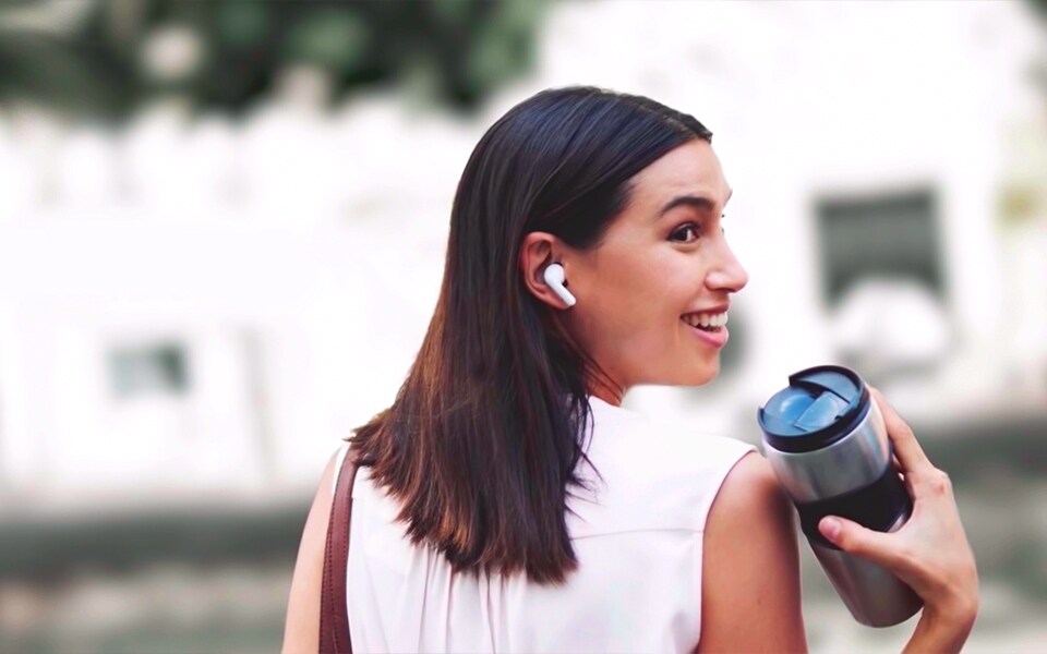 A woman looking back and smiling whilst carrying a cup and wearing earphones