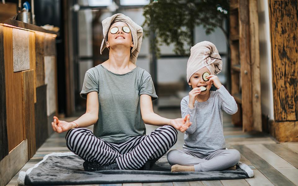 A woman practising meditation whilst wearing matching head towels and cucumber slices as the child sitting next to her.