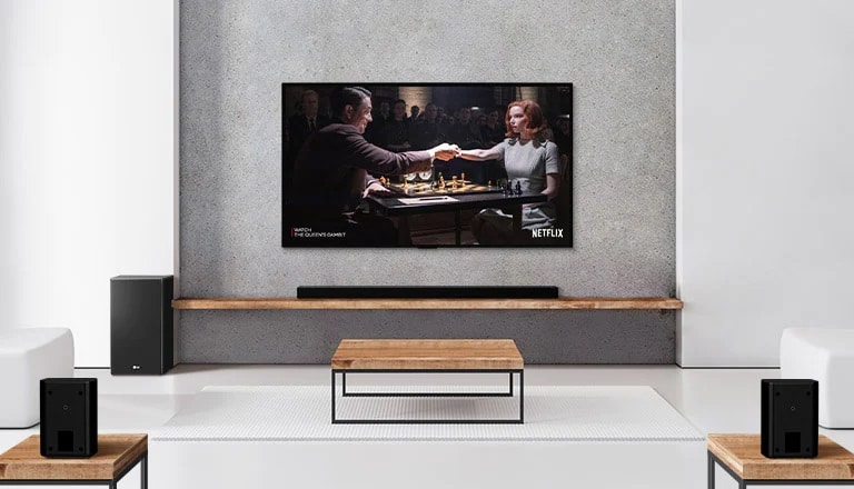 Un set de 2 difuzoare spate, un subwoofer și o bară de sunet și televizorul sunt într-o cameră de zi albă.  O femeie și un bărbat joacă șah pe ecranul televizorului.
