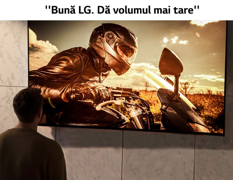 Perspectivă din spatele unui bărbat care se uită la televizorul de pe peretele din sufragerie. Pe ecran se vede o scenă cu o motocicletă dintr-un film.