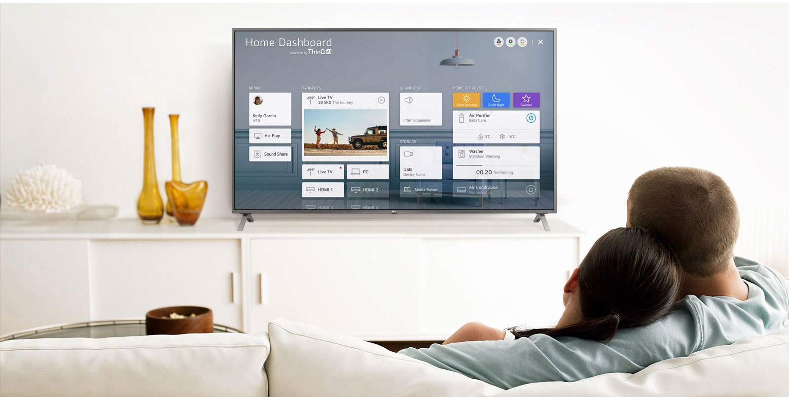 Un bărbat și o femeie care stau pe o canapea în camera de zi, cu Home Dashboard pe ecranul TV.