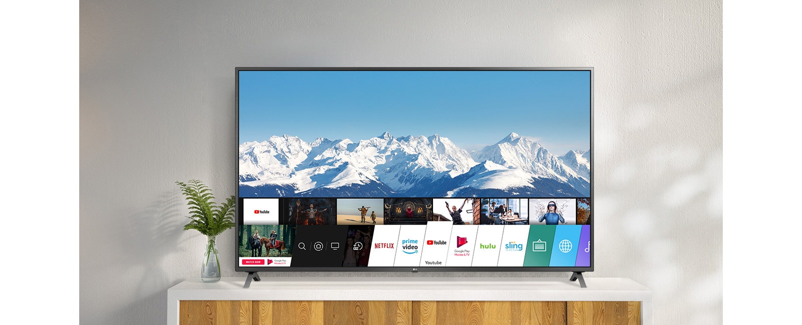 Televizor amplasat pe un suport alb, pe un perete alb. Ecranul TV prezintă ecranul principal cu webOS.