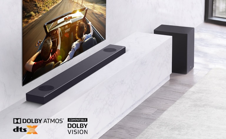 Televizorul este amplasat pe perete, soundbarul LG este instalat dedesubt, pe un raft de marmură albă, având în dreapta subwooferul. Pe televizor se afișează un cuplu într-o mașină. 