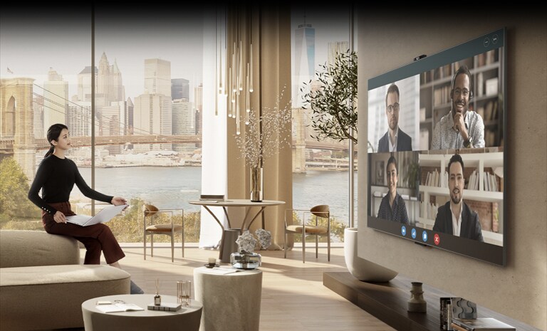 O femeie stă pe o canapea într-o cameră de zi spațioasă. Aceasta ține în mână un laptop în timp ce se află în fața unui ecran mare de pe peretele opus, ceea ce indică faptul că se află într-o videoconferință.