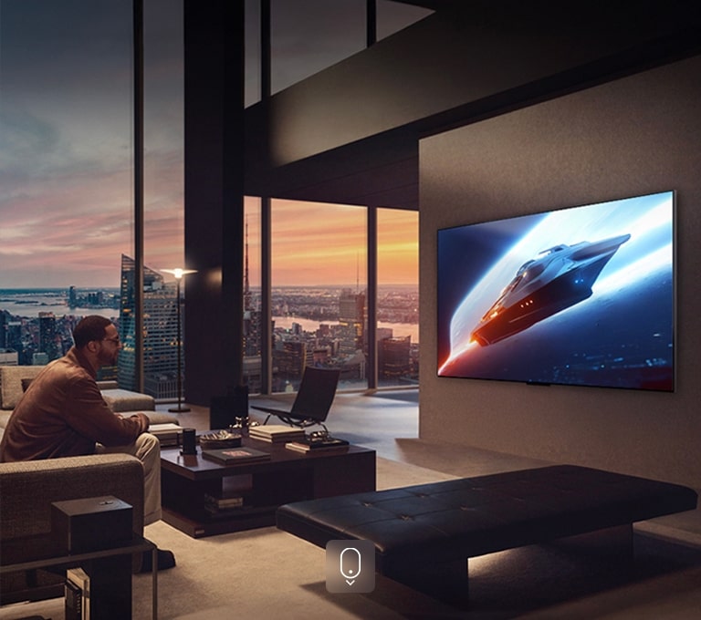 Sufragerie elegantă într-un apartament tip penthouse. Orizontul orașului este vizibil pe fereastră. Un bărbat stă pe canapea, urmărim conținut pe un TV montat pe perete. 