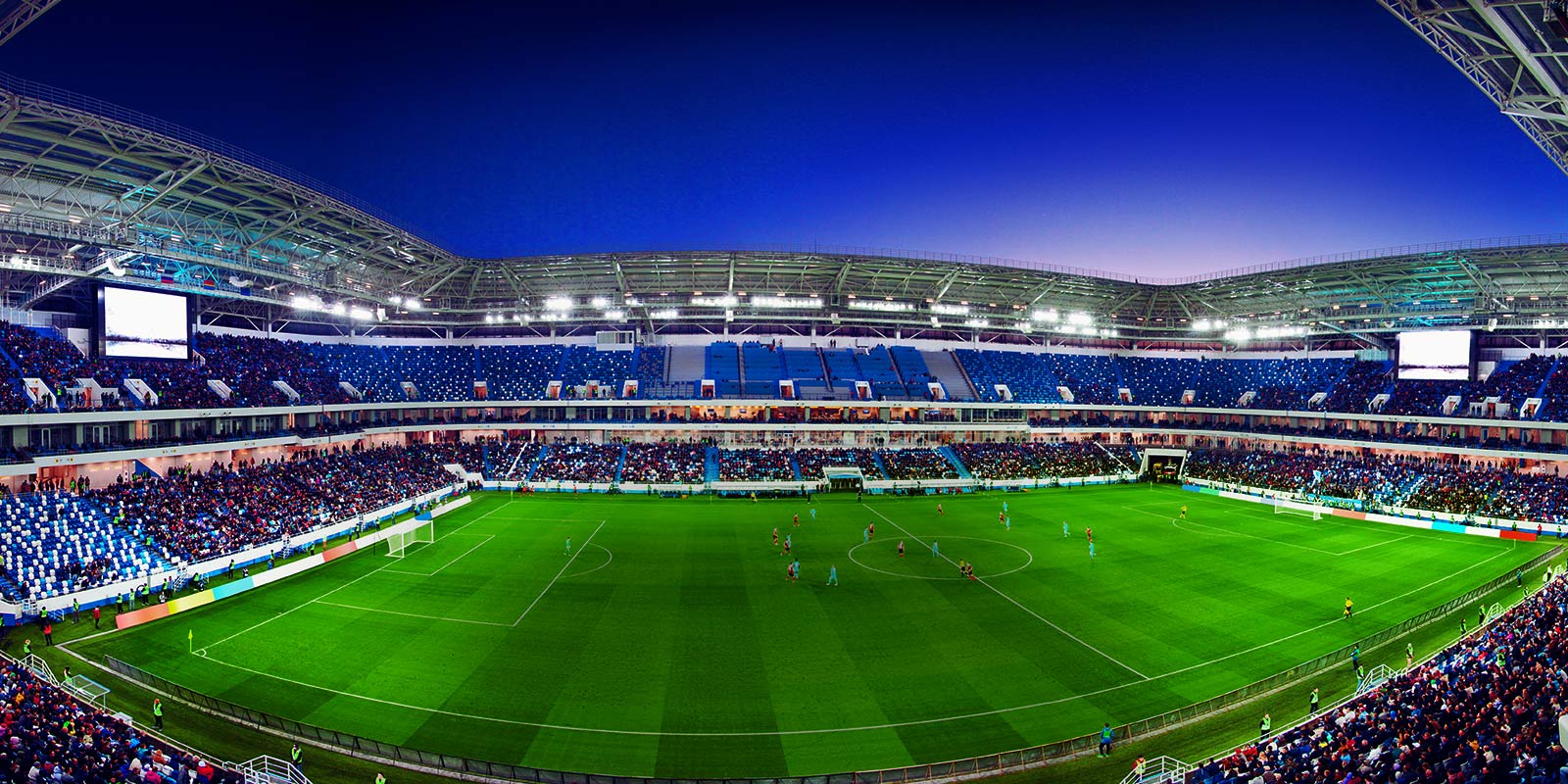 O vedere cu unghi larg a unui stadion de fotbal plin cu spectatori și cu un meci în desfășurare.