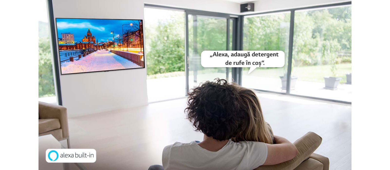 Cuplu stând pe o canapea, cerându-i aplicației Amazon Alexa să adauge detergent de rufe în coșul de cumpărături