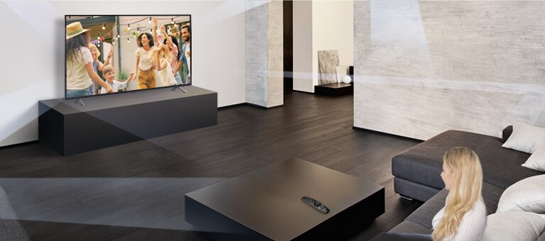 O doamnă într-o sufragerie spațioasă și simplistă, privind la televizor oameni care dansează.