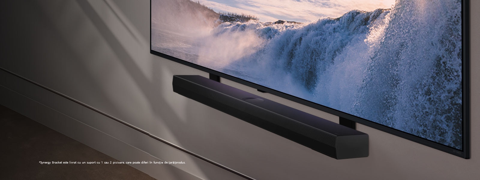 LG TV și soundbar sunt așezate într-o poziție înclinată, montate pe un perete. Pe televizor este afișat un prim plan al unei vaste cascade, iar lumina blândă a soarelui se îndreaptă peste perete, televizor și soundbar. O declinare a răspunderii transmite: „Suportul Synergy vine însoțit de un suport cu 1 pol sau 2 poli, care pot diferi în funcție de țară/produs.”