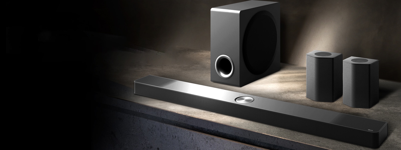 LG soundbar, difuzoare spate și subwoofer așezate într-o perspectivă înclinată pe un raft maro din lemn, într-o cameră neagră, învăluită în întuneric, cu lumină doar pe sistemul audio.