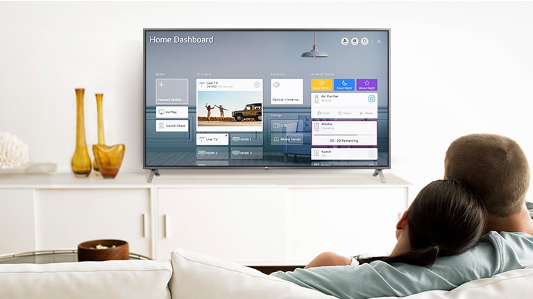 Un bărbat și o femeie care stau pe o canapea în camera de zi, cu Home Dashboard pe ecranul TV.