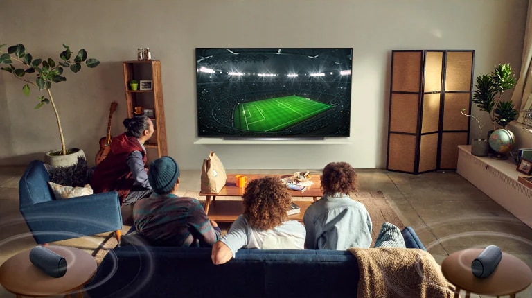 Oameni care urmăresc un joc sportiv la televizor în camera de zi, cu difuzoare Bluetooth în spate
