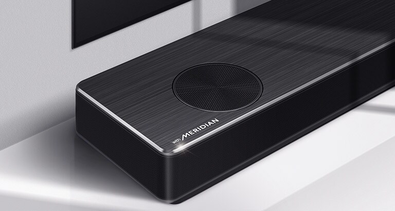 Krupan kadar leve strane LG Soundbar zvučnika sa logotipom Meridian prikazanim u donjem levom uglu proizvoda. Donja leva strana televizora je takođe vidljiva.