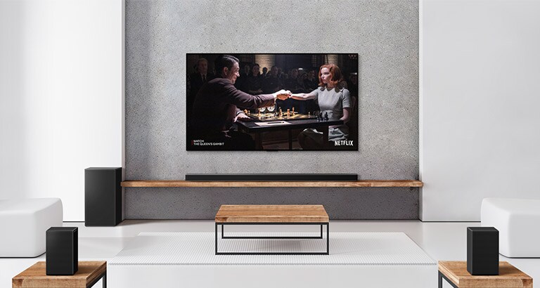 Komplet 2 zadnja zvučnika, niskotonac i Soundbar zvučnik i televizor nalaze se u beloj dnevnoj sobi. Žena i muškarac igraju šah na TV ekranu.