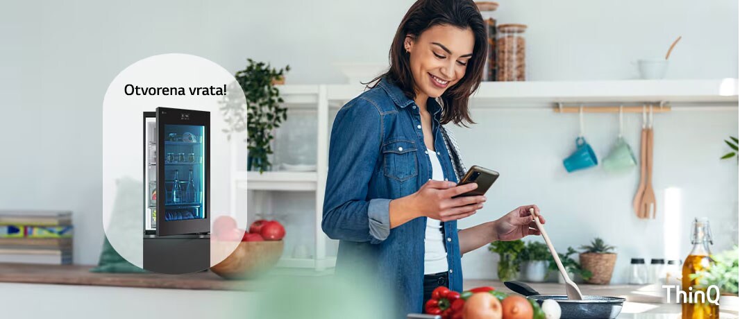 Slika žene koja prima obaveštenje na svoj pametni telefon iz aplikacije LG ThinQ o tome da su vrata frižidera ostavljena otvorena dok ona kuva.