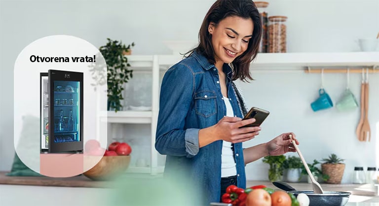 Slika žene koja prima obaveštenje na svoj pametni telefon iz aplikacije LG ThinQ o tome da su vrata frižidera ostavljena otvorena dok ona kuva.