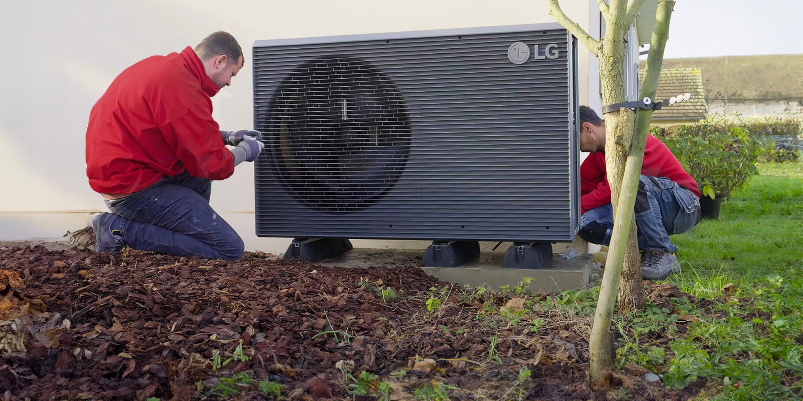 Dva instalatera u crvenim radnim uniformama instaliraju LG toplotnu pumpu vazduh/voda THERMA V R290 Monoblok u dvorištu kuće.