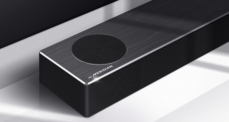 Krupan kadar leve strane LG Soundbar zvučnika sa logotipom Meridian prikazanim u donjem levom uglu proizvoda. Donja leva strana televizora je takođe vidljiva.
