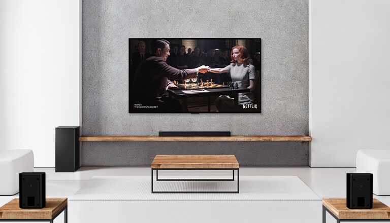 Komplet 2 zadnja zvučnika, niskotonac i Soundbar zvučnik i televizor nalaze se u beloj dnevnoj sobi. Žena i muškarac igraju šah na TV ekranu.