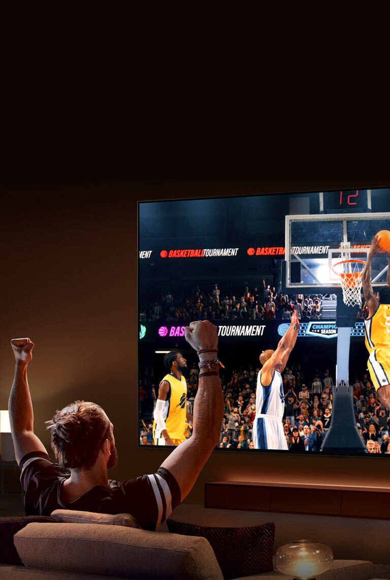Muškarac sedi na sofi ispred izuzetno velikog LG TV-a na kom je košarkaška utakmica, sa podignutim rukama kao da navija.
