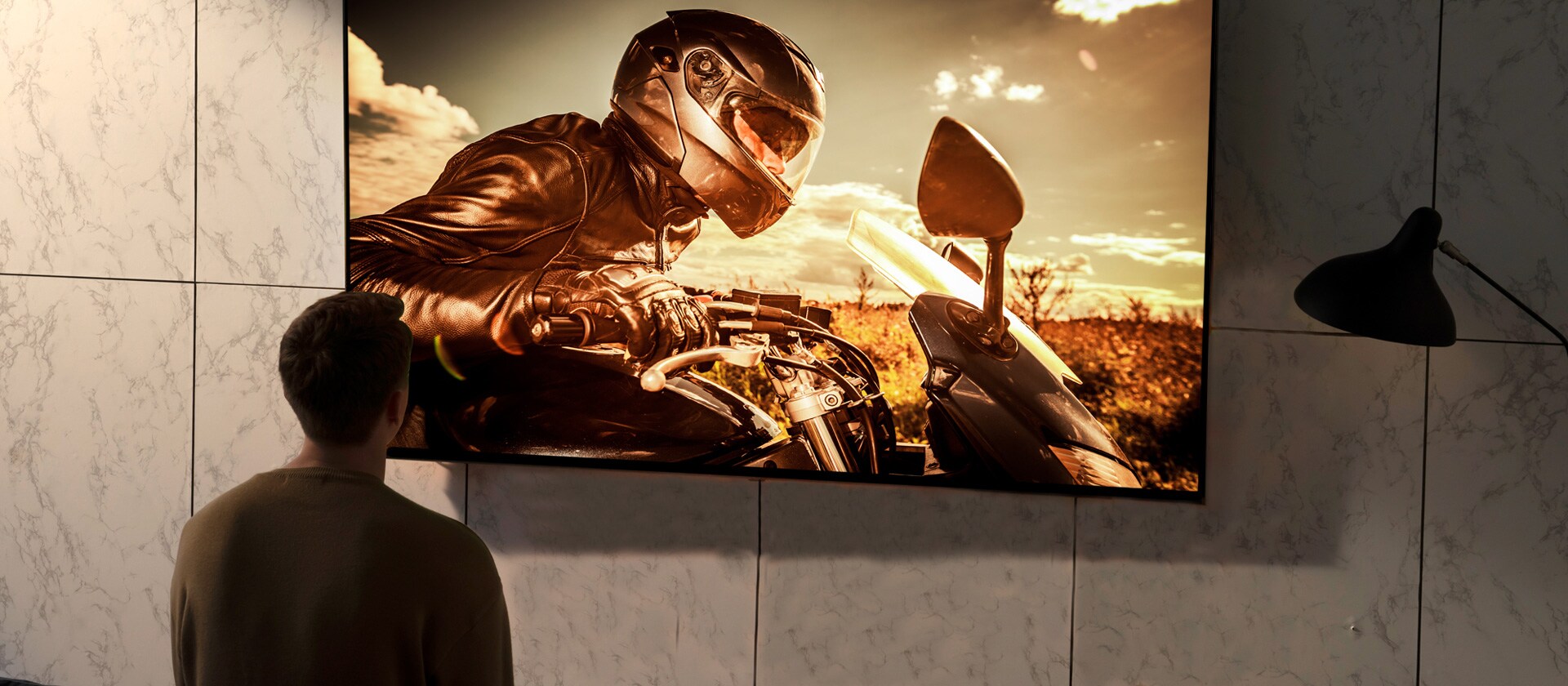 Prikaz čoveka otpozadi kako gleda televizor na zidu u dnevnoj sobi. Na ekranu je scena iz filma sa motociklom.