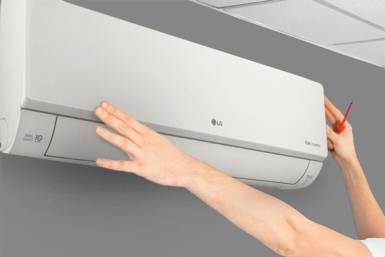 На стене установлен кондиционер, вид с боку. Две руки тянутся вверх, одна держит инструмент
