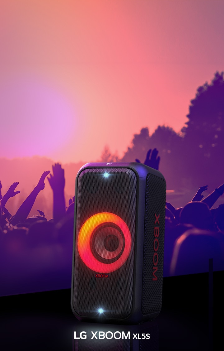 LG XBOOM XL5T стоит на сцене с включенной подсветкой с красно-оранжевым градиентом. За сценой люди наслаждаются музыкой.