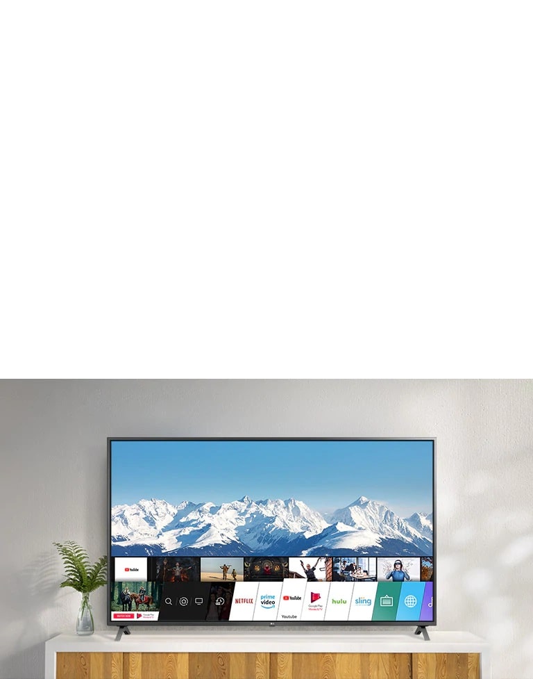 Телевизор стоит на белой тумбе на фоне белой стены. На экране показан рабочий стол с webOS.